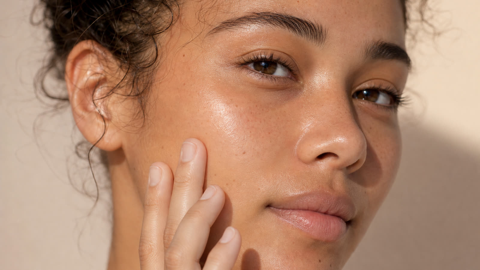 Close-up of a young woman with clear, healthy, glowing skin gently touching her cheek in soft daylight