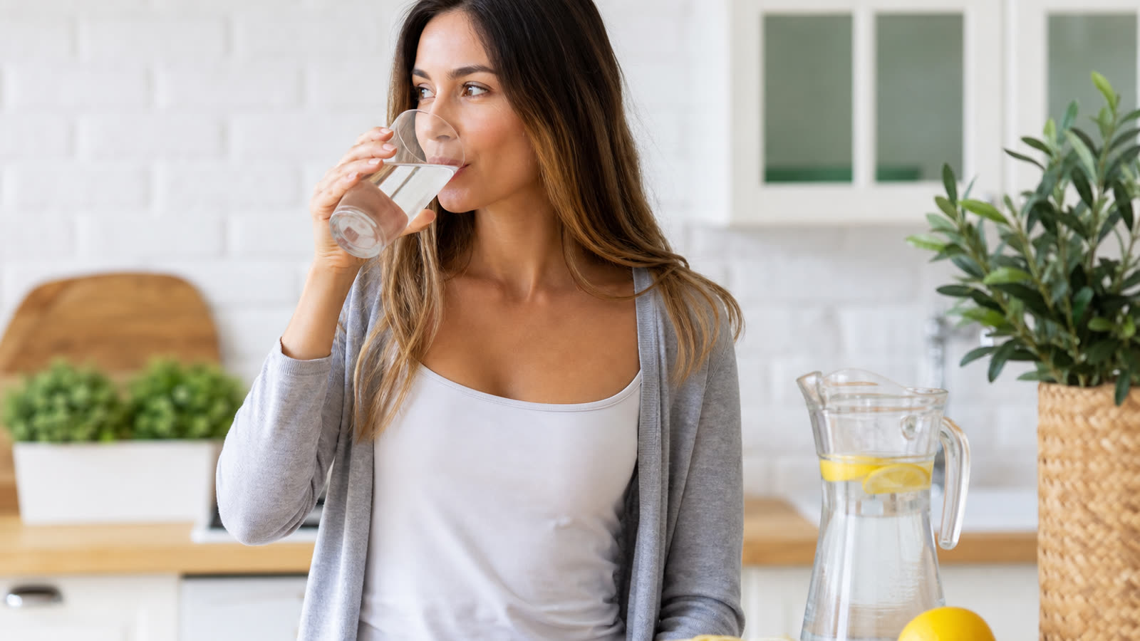 Woman drinking a tall glass of water in a bright kitchen with a water pitcher and lemon slices