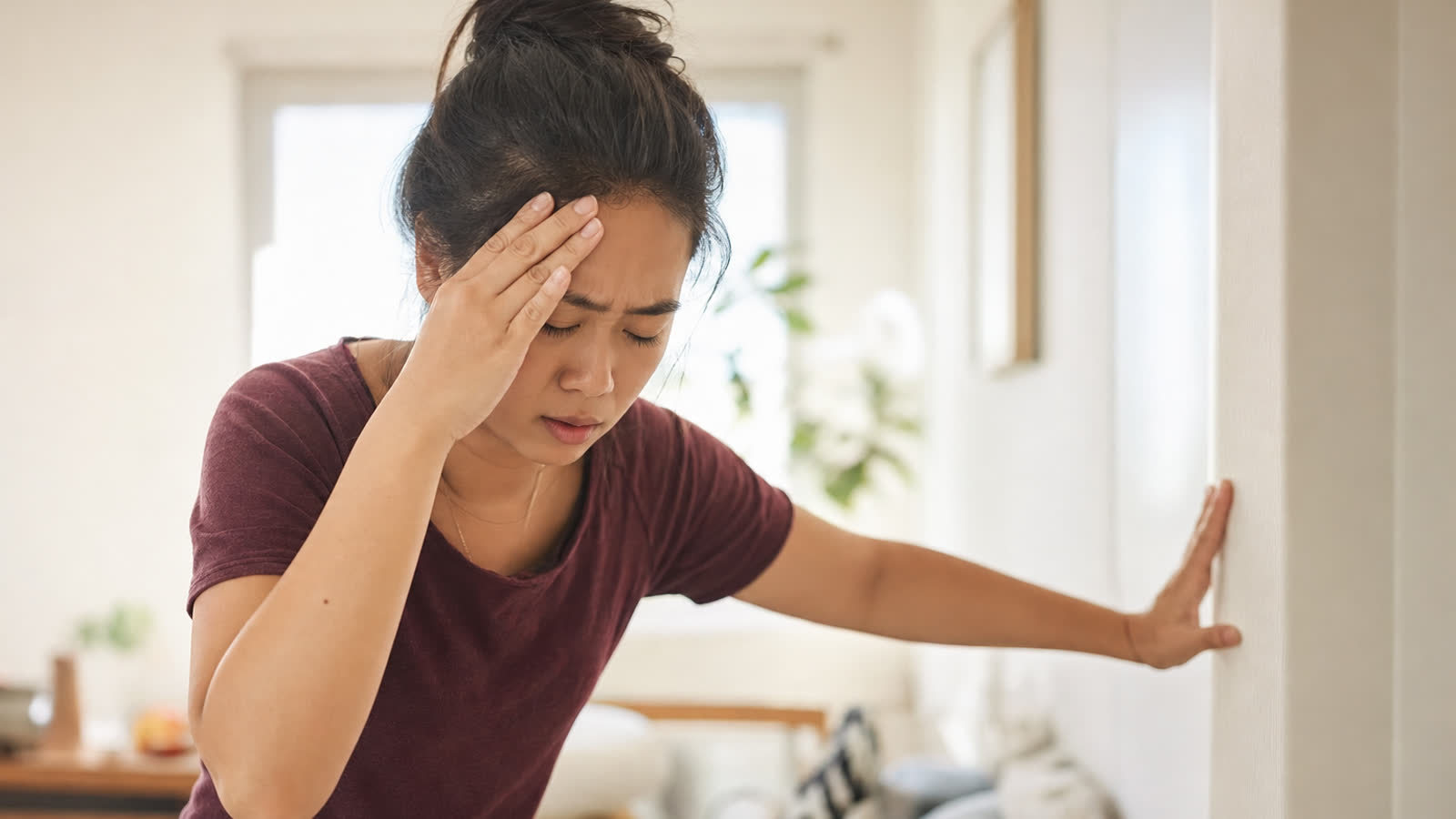 Person pressing a hand to their forehead feeling dizzy and lightheaded after standing up, looking unsteady in a bright room