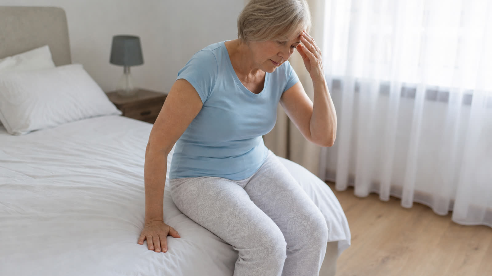 Older adult standing up slowly from the edge of a bed with a hand on the mattress for balance, illustrating orthostatic hypotension