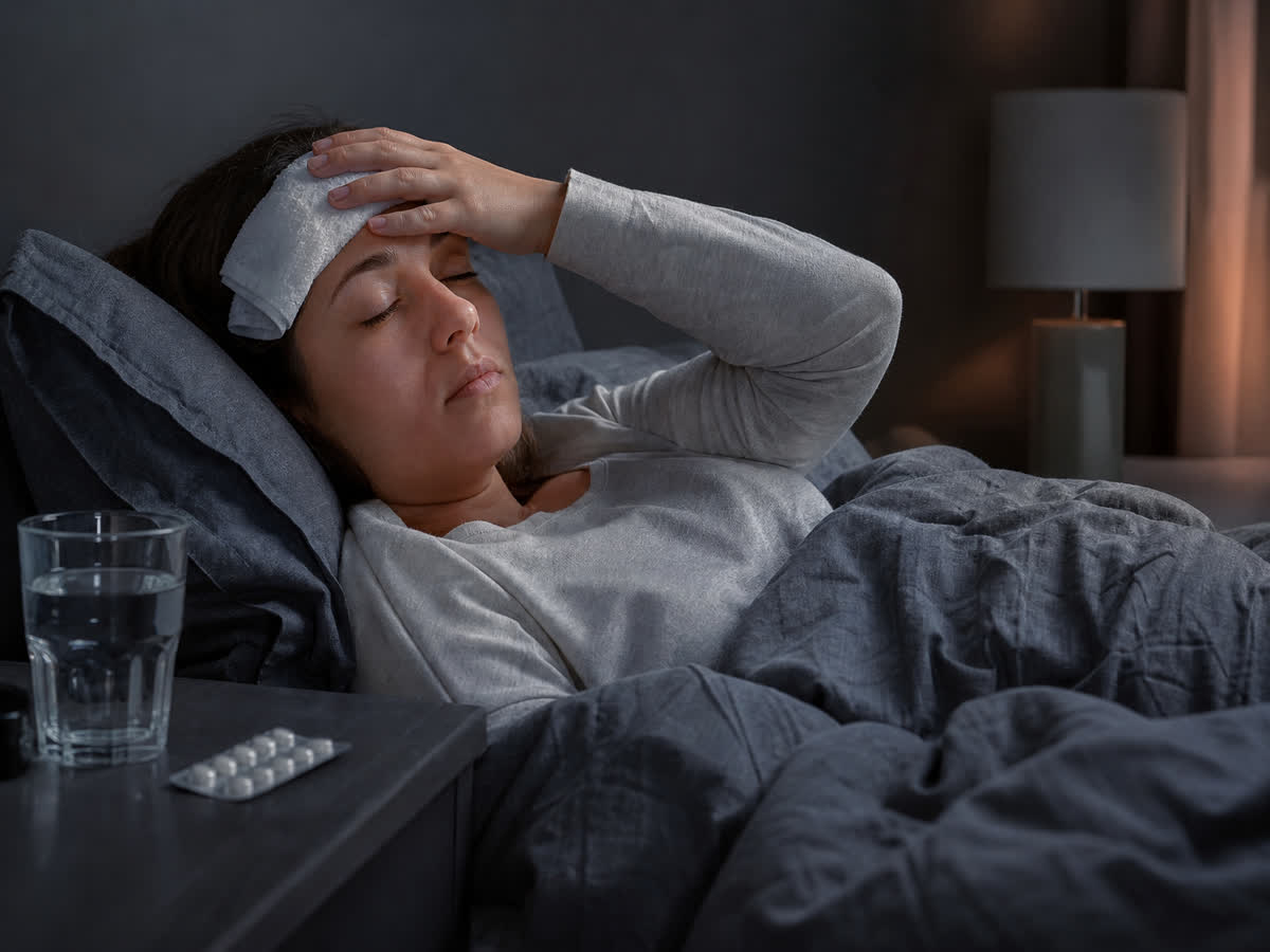 Person resting in a dark quiet bedroom with a cool cloth on the forehead, water and pills on the nightstand
