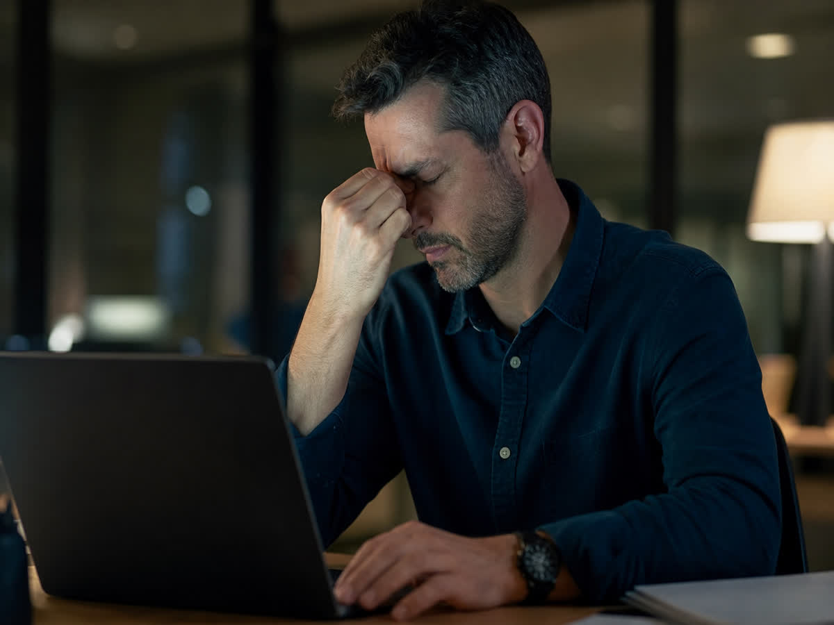 Man at a desk pinching the bridge of his nose with eyes closed from a screen-strain tension headache
