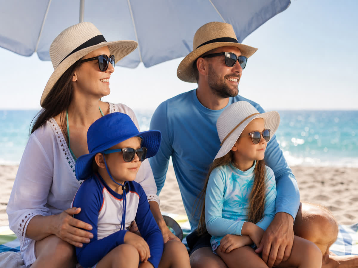 A family wearing wide-brimmed hats and sunglasses relaxing under a beach umbrella for shade on a sunny day
